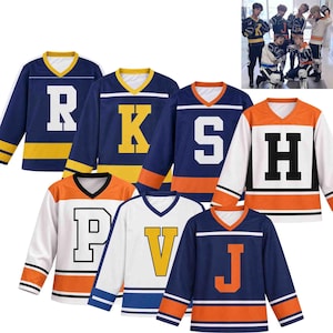 Op de afbeelding: Een collectie van zes sportshirts met lange mouwen en V-hals in diverse kleurcombinaties, waaronder marineblauw, wit en oranje. Elk shirt is voorzien van een grote blokletter op de borst, waaronder R, K, S, H, P, V en J, met gestreepte mouwboorden.