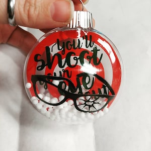 May include: A red and white Christmas bauble with the text "You'll shoot your eye out" and a black silhouette of a pair of glasses. The bauble is filled with white polystyrene balls.