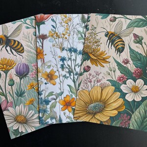 May include: Three notebooks with floral and bee designs. The covers feature illustrations of yellow and white daisies, purple flowers, and bees. The background is a light beige color with green leaves and other wildflowers.
