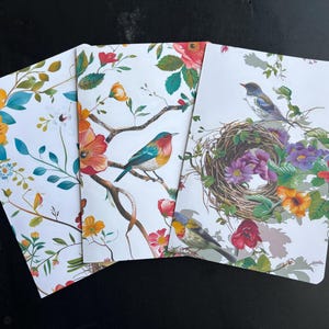May include: Three notebooks with floral and bird designs. The covers feature colorful illustrations of birds perched on branches, surrounded by flowers in shades of yellow, orange, red, and purple, set against a white background.