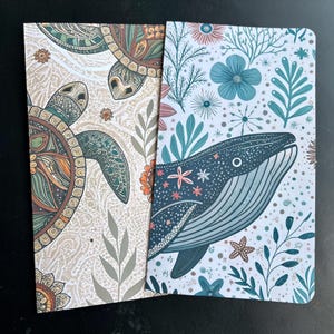 May include: Two notebooks with decorative covers. One features a detailed turtle design in shades of brown, green, and orange against a cream background. The other notebook has a whale illustration with blue and teal accents, surrounded by floral and botanical elements.