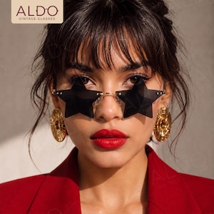 May include: Star-shaped sunglasses with dark tinted lenses and a thin gold-tone metal frame. The glasses feature a bridge and nose pads, and the text ALDO VINTAGE GLASSES is visible in the upper left corner of the frame.
