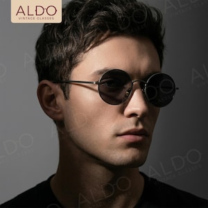 May include: Round sunglasses with thin dark metal frames and dark tinted lenses. The glasses feature a minimalist design with a slim bridge and temples. The logo ALDO VINTAGE GLASSES is visible in the top left corner of the image frame.