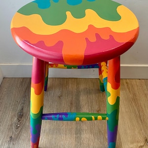 May include: A round wooden stool painted with a vibrant rainbow design. The seat features a flowing pattern of red, orange, yellow, green, and blue. The legs and supports are also painted in a rainbow gradient, creating a colorful accent piece.