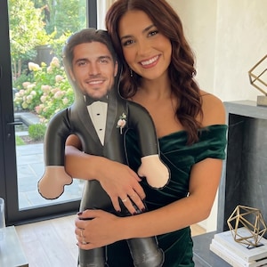May include: A woman holding an inflatable doll of a man in a black tuxedo with a bow tie and a pink buttonhole. The doll has a photo of a man's face. The woman wears a dark green off-the-shoulder dress.