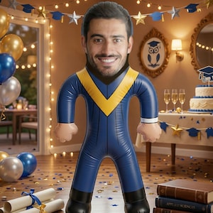 May include: An inflatable graduation figure with a smiling face, wearing a blue and yellow graduation gown. The figure stands in a room decorated for a graduation party, with balloons, a cake, and books.