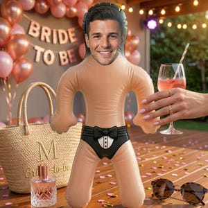 Könnte beinhalten: Aufblasbare Puppe mit männlichem Gesicht, trägt schwarze Unterwäsche mit Fliege und Smoking-Design. Eine Strohtasche, eine Parfümflasche und ein Glas rosa Getränk stehen auf einem Holztisch. Rosa Luftballons und ein "Bride to Be" Banner sind im Hintergrund.