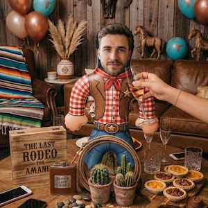 May include: An inflatable cowboy doll with a face, wearing a red and white checked shirt, brown waistcoat, and blue jeans. A wooden box with the text "THE LAST RODEO FOR AMANI" is next to a leather hip flask. The scene includes balloons, cacti, and pastries.