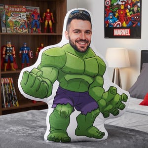 Peut inclure: Un grand coussin personnalisé en forme de Hulk, avec le visage d'un homme souriant. Le coussin est principalement vert avec un short violet. L'arrière-plan comprend une affiche Marvel et des figurines.