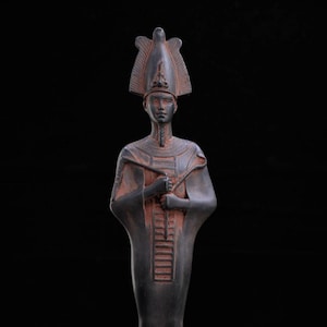 Puede incluir: Estatua de bronce oscuro de un faraón egipcio, con una alta tocado y vestimenta ceremonial. La estatua tiene un rostro detallado y sostiene un bastón. El fondo es negro sólido.