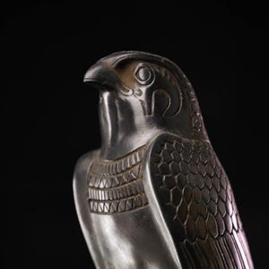 Op de afbeelding: Een close-up van een zilverkleurige valkensculptuur. De kop van de vogel is gedraaid en toont gedetailleerde kenmerken van het oog, de snavel en de veren. De sculptuur heeft een getextureerd oppervlak en staat tegen een zwarte achtergrond.