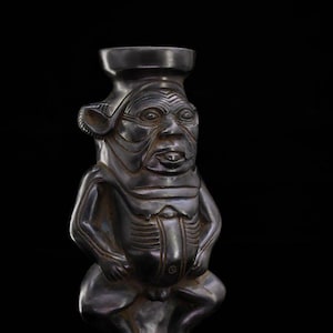 Może przedstawiać: Ciemna metalowa rzeźba siedzącej postaci humanoidalnej ze szczegółową twarzą, dużymi uszami i cylindrycznym nakryciem głowy. Ciało postaci ma wytrawione linie i zaokrąglony brzuch na czarnym tle.