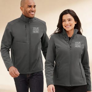 May include: Two charcoal gray softshell jackets with the text "YOUR DESIGN HERE" printed on the chest. The jackets have a zip-up front, a stand-up collar, and multiple pockets. The jackets are worn by two people.