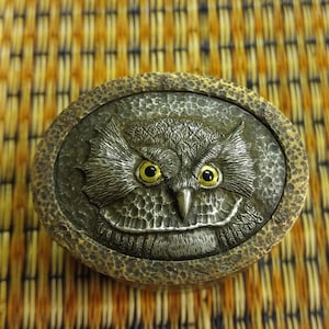 May include: A silver owl with yellow eyes is embossed on a round, brown metal box.