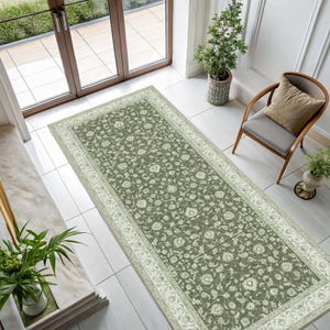 May include: A rectangular area rug featuring an olive green field with a dense, intricate floral and vine pattern in cream. The rug is framed by a wide, ornate cream-colored border with repeating botanical motifs, creating a balanced design.