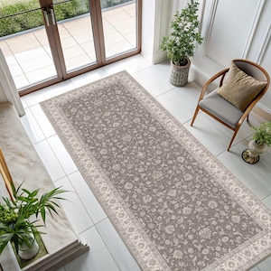 May include: A rectangular area rug featuring a taupe-colored field with an intricate, repeating floral pattern in a lighter cream shade. The rug is framed by a wide, decorative border that mirrors the floral motif, creating a cohesive design.