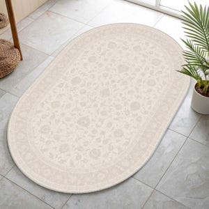 May include: An oval-shaped area rug featuring a light beige background with a subtle, repeating floral pattern in a slightly darker cream tone. The rug has a decorative border with intricate botanical motifs and a clean, finished edge throughout.