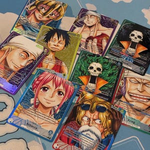 May include: A collection of colorful trading cards featuring anime-style illustrations of various characters. Each card has a unique design with character names and the number "5000" printed on them. The cards are arranged on a light blue surface.