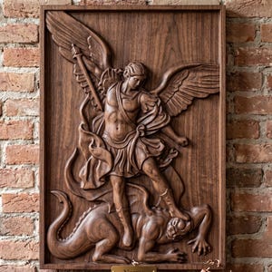 May include: A rectangular dark brown wooden wall plaque featuring a detailed relief carving of a winged figure in armor holding a sword and standing over a dragon. The carving is set within a simple wooden frame with a small gold plaque at the bottom.