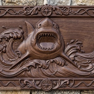 May include: A rectangular dark brown wooden wall plaque featuring a detailed relief carving of a shark emerging from ocean waves. The frame is decorated with nautical motifs including anchors, seashells, and compass roses. Text reads Digital Download - STL.