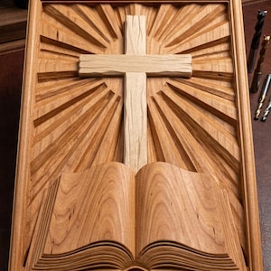 May include: A rectangular wooden wall plaque featuring a carved open book at the bottom and a central cross above it, surrounded by radiating sunburst lines. The text Digital Download - STL is engraved in the bottom right corner of the frame.