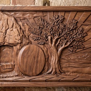 May include: A rectangular wooden wall plaque featuring a detailed relief carving of a tomb entrance with a large circular stone rolled to the side, a textured rocky cliff face, a leafy tree, and sun rays emanating across the background sky.