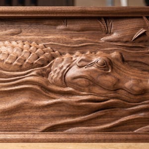 May include: A rectangular dark brown wooden wall plaque featuring a detailed relief carving of an alligator swimming through water. The alligator is shown in profile with textured scales, and the scene includes carved reeds in the upper right corner.