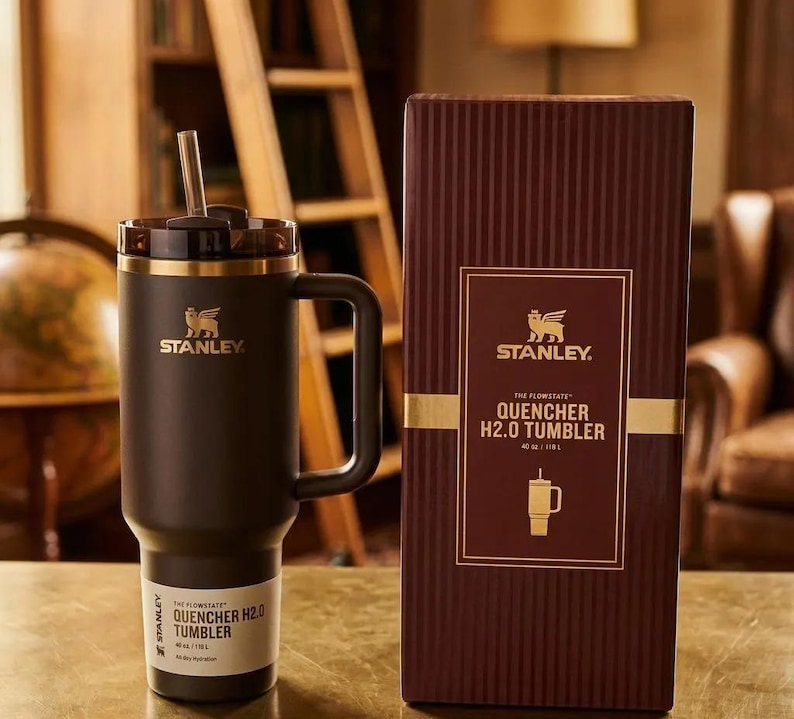 May include: A black Stanley Quencher H2.0 tumbler with a handle and a straw, next to its brown box. The box features the Stanley logo and product details. The tumbler is 40 oz / 1.18 L.