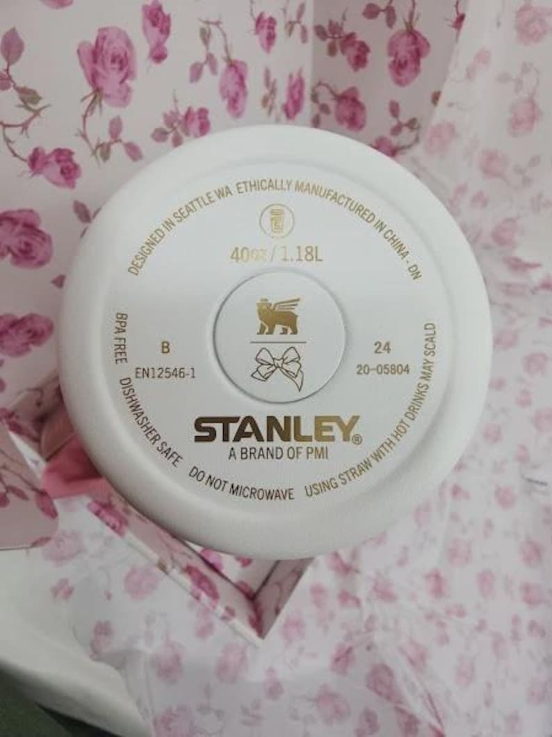 May include: White Stanley brand tumbler lid with gold accents. The lid features the Stanley logo, a lion emblem, and text indicating a 40 oz / 1.18L capacity. The lid is dishwasher safe and designed in Seattle, WA.