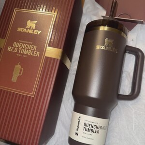 May include: A brown Stanley Quencher H2.0 tumbler with a handle and straw, next to its matching box. The box is brown with gold accents and the Stanley logo. The tumbler holds 40 oz.