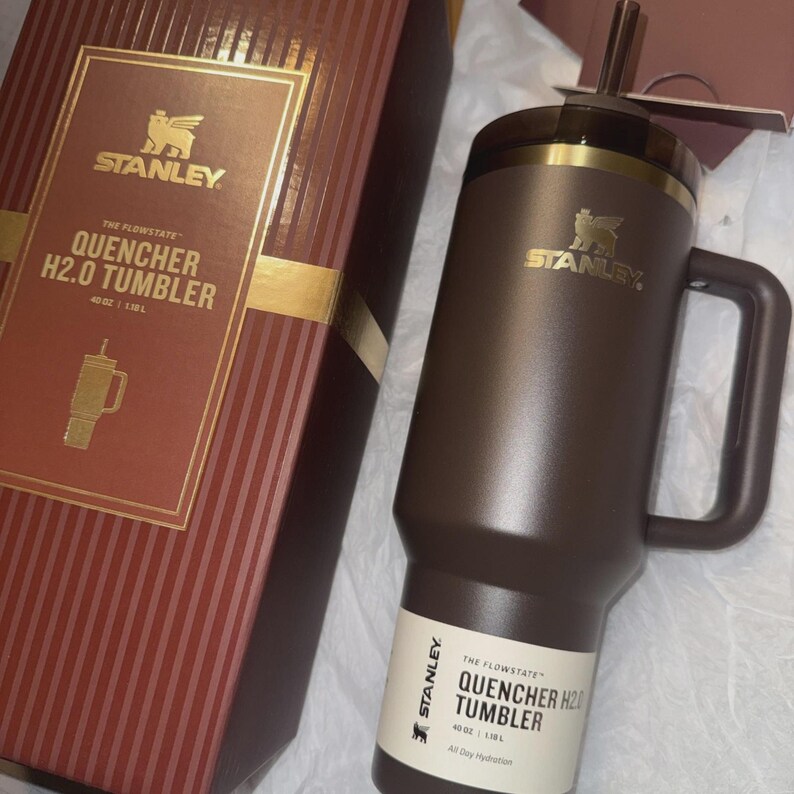 May include: A brown Stanley Quencher H2.0 tumbler with a handle and straw, next to its matching box. The box is brown with gold accents and the Stanley logo. The tumbler holds 40 oz.