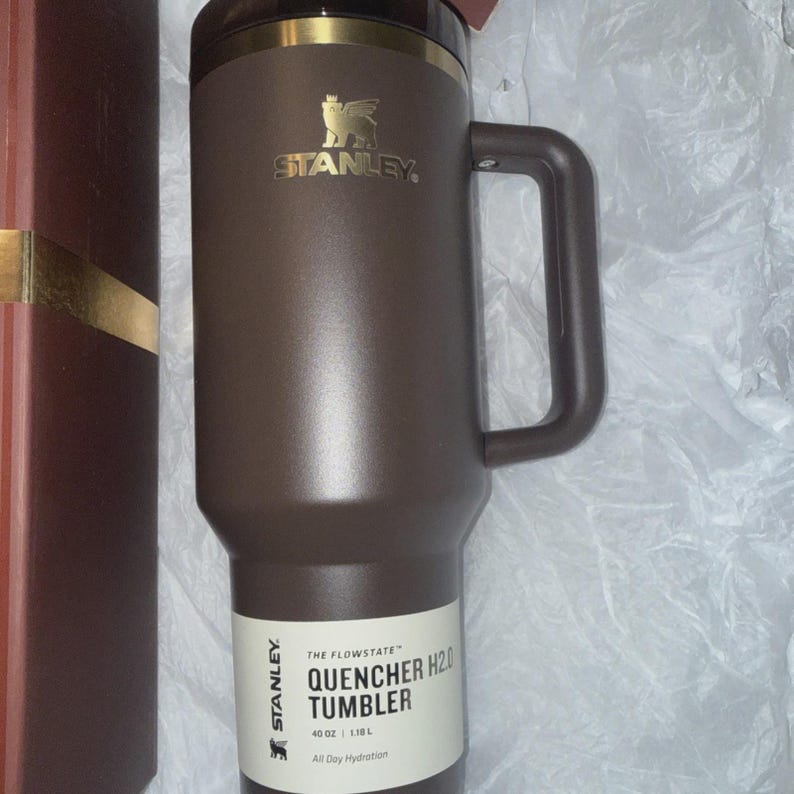 May include: A brown Stanley Quencher H2.0 tumbler with a handle. The tumbler has a gold-colored rim and the Stanley logo. The label reads "QUENCHER H2.0 TUMBLER" and "40 OZ | 1.18 L". The tumbler is designed for all-day hydration.