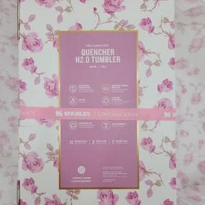 May include: A box with a floral rose pattern in pink and white. The box features a pink rectangle with the text "QUENCHER H2.0 TUMBLER" and the Stanley and Loveshackfancy logos. A pink ribbon wraps around the box.