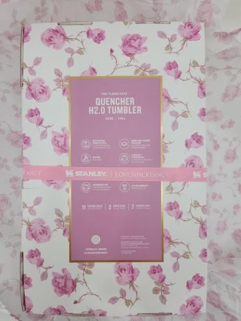 May include: A box with a floral rose pattern in pink and white. The box features a pink rectangle with the text "QUENCHER H2.0 TUMBLER" and the Stanley and Loveshackfancy logos. A pink ribbon wraps around the box.