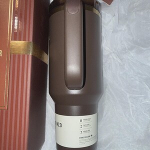 May include: A brown insulated tumbler with a handle and a gold-colored rim. The tumbler has a label with the text "H2.0" and information about how long it keeps drinks cold and hot. The tumbler is next to a brown and red striped box.
