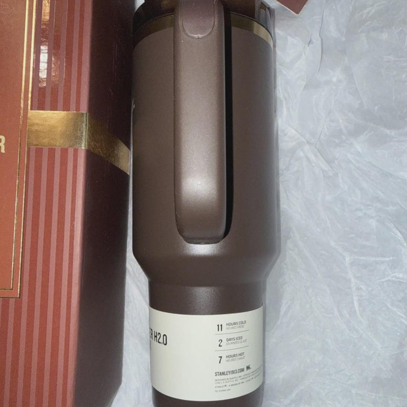 May include: A brown insulated tumbler with a handle and a gold-colored rim. The tumbler has a label with the text "H2.0" and information about how long it keeps drinks cold and hot. The tumbler is next to a brown and red striped box.