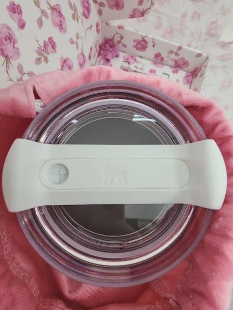 May include: A clear, round container with a white handle. The container is set on a pink surface, with a floral patterned background. The handle has a button and a logo. The container appears to be a food storage item.