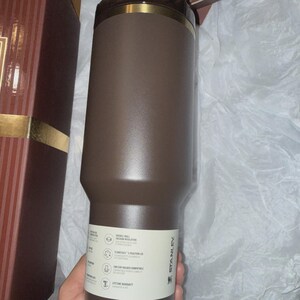 May include: A brown Stanley tumbler with a gold rim and a white label. The tumbler has a double-wall vacuum insulation and is car cup holder compatible. The tumbler is in a gift box.