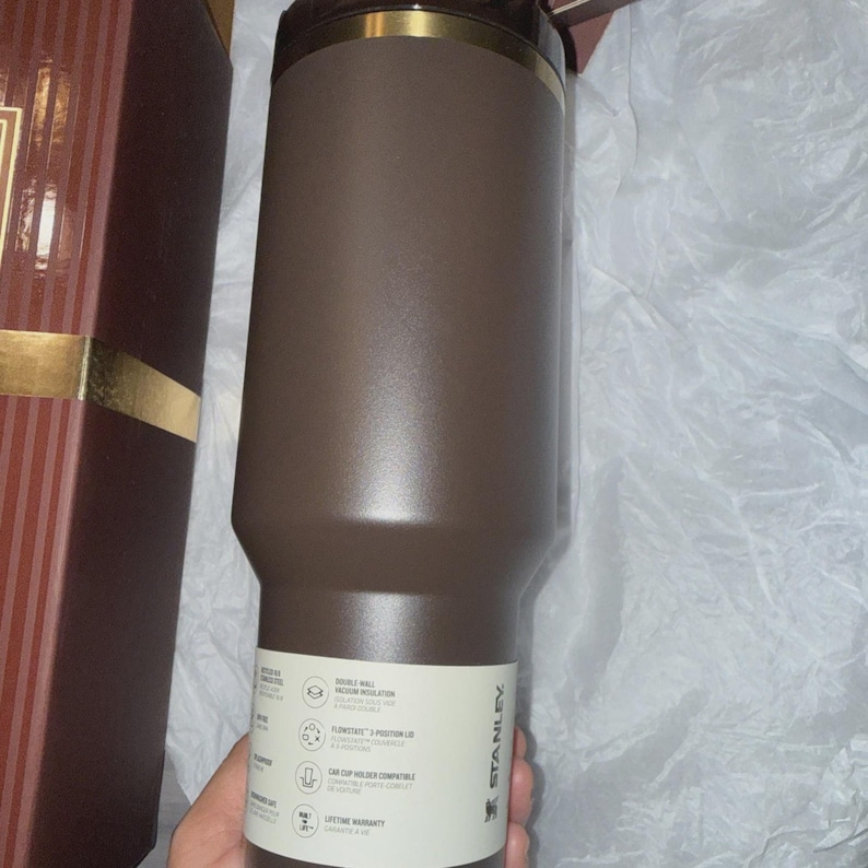 May include: A brown Stanley tumbler with a gold rim and a white label. The tumbler has a double-wall vacuum insulation and is car cup holder compatible. The tumbler is in a gift box.
