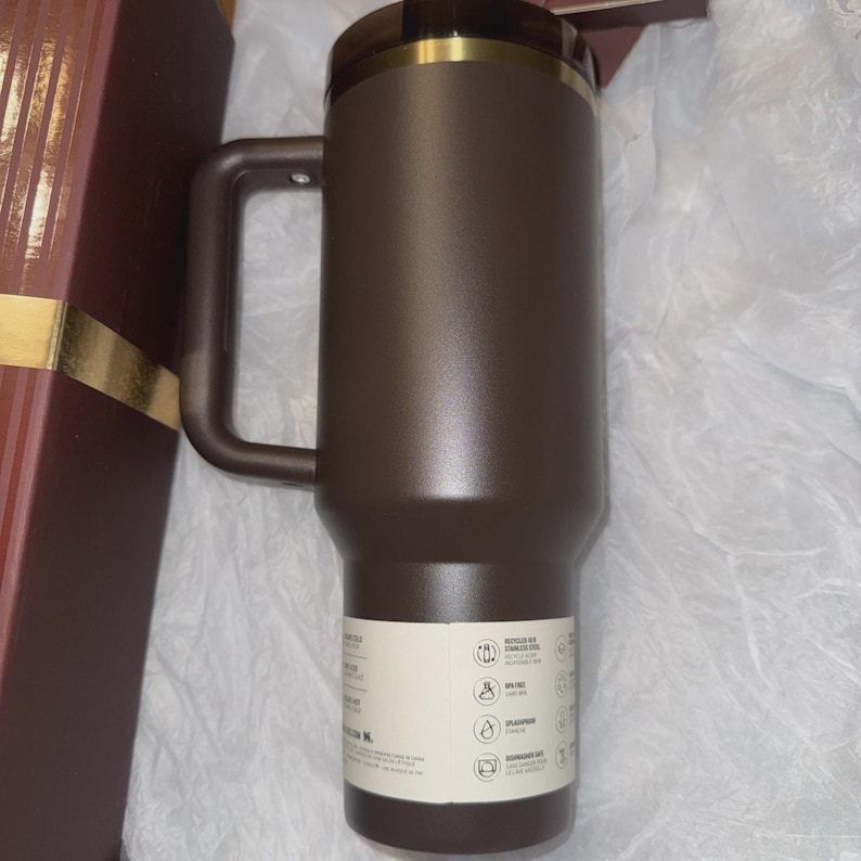 May include: A brown insulated travel mug with a handle and a dark brown lid with a gold rim. The mug has a matte finish and a white label with icons and text. The mug is in a gift box.