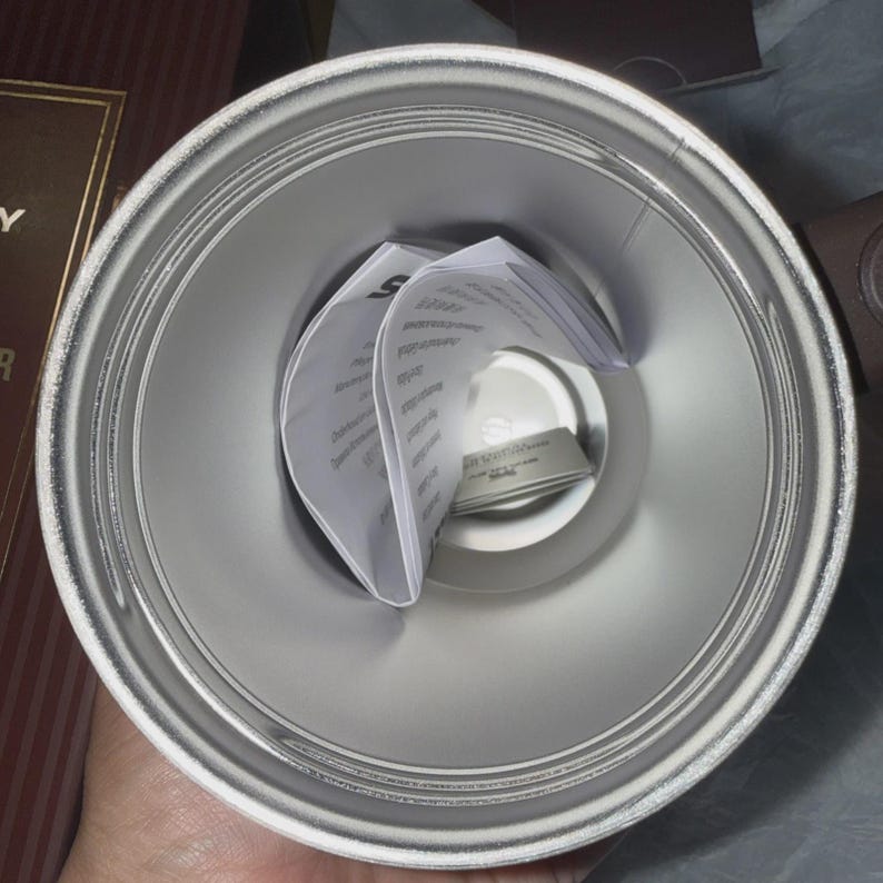May include: A silver-colored insulated tumbler, viewed from above, with a wide opening and a narrower base. Inside, folded white paper inserts are visible. The tumbler appears to be new and unused.