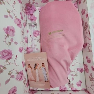 May include: A pink Stanley Loveshackfancy insulated container inside a floral box. The box is decorated with pink roses on a white background. A small booklet with women in dresses is visible.