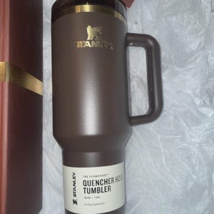 May include: A brown Stanley Quencher H2.0 tumbler with a handle. The tumbler has a gold-colored rim and the Stanley logo. The label reads "QUENCHER H2.0 TUMBLER" and "40 OZ | 1.18 L". The tumbler is designed for all-day hydration.