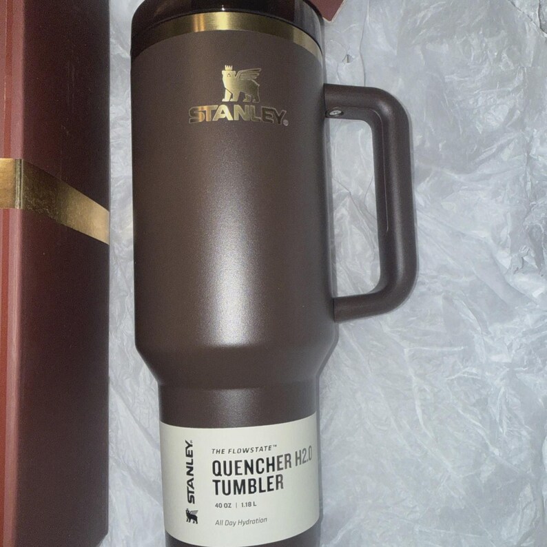 May include: A brown Stanley Quencher H2.0 tumbler with a handle. The tumbler has a gold-colored rim and the Stanley logo. The label reads "QUENCHER H2.0 TUMBLER" and "40 OZ | 1.18 L". The tumbler is designed for all-day hydration.