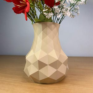 Könnte beinhalten: Eine beige geometrische Vase mit roten und weißen Blumen. Die Vase hat ein facettiertes Design mit rautenförmigen Paneelen. Die Blumen umfassen rote Lilien und kleine weiße Blüten, wodurch ein dekoratives Blumenarrangement entsteht.