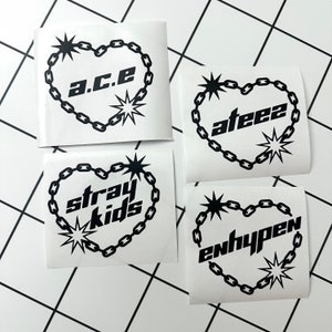 May include: Four white square stickers with black chain heart designs and band names. The stickers feature the names "a.c.e", "ateez", "stray kids", and "enhypen".