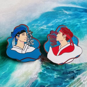 May include: Two enamel pins depicting two figures in traditional clothing. The figure on the left is wearing blue and white clothing with a blue headband and is surrounded by white flowers. The figure on the right is wearing red and white clothing with a red headband and is surrounded by red flowers. Both figures are looking at each other.