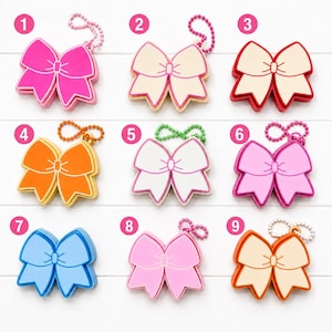 May include: A collection of nine bow-shaped keychains, each numbered one through nine. The bows feature various solid colors including hot pink, cream, red, orange, white, light pink, blue, pastel pink, and peach, all with attached ball chains.