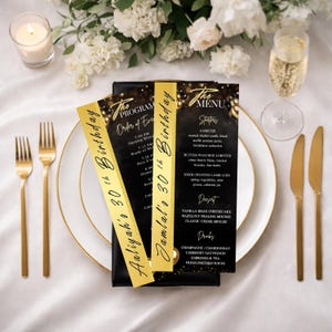 May include: Elegant table setting with gold cutlery, white plates, and champagne glasses. Two menu cards and two program cards with gold accents and the text "Aaliyah's 30th Birthday" and "Jamal's 30th Birthday" are displayed. White flowers and a candle complete the festive scene.