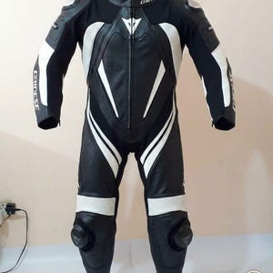 K&ouml;nnte beinhalten: Schwarzer und wei&szlig;er Motorrad-Rennanzug aus Leder mit dem Markennamen "Dainese" auf Brust und Armen. Der Anzug hat wei&szlig;e Streifen und Polsterungen an Schultern, Armen und Beinen. Knieschleifer sind sichtbar.